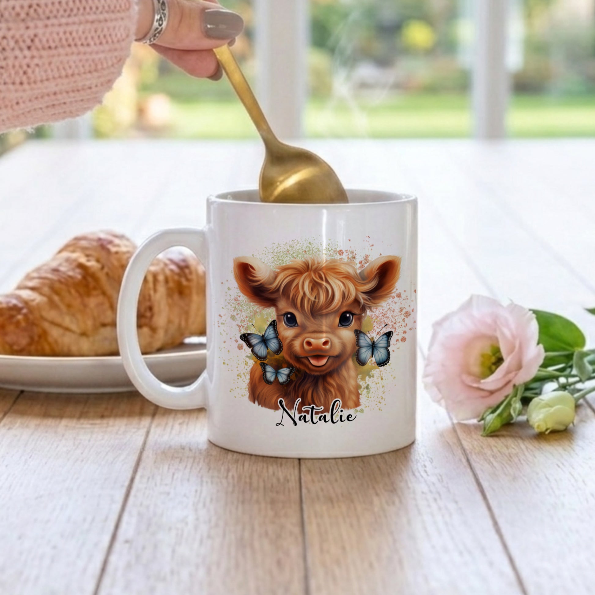 Mug with a cow design and name 'Natalie', being stirred with a spoon, on a table with a croissant and flowers.