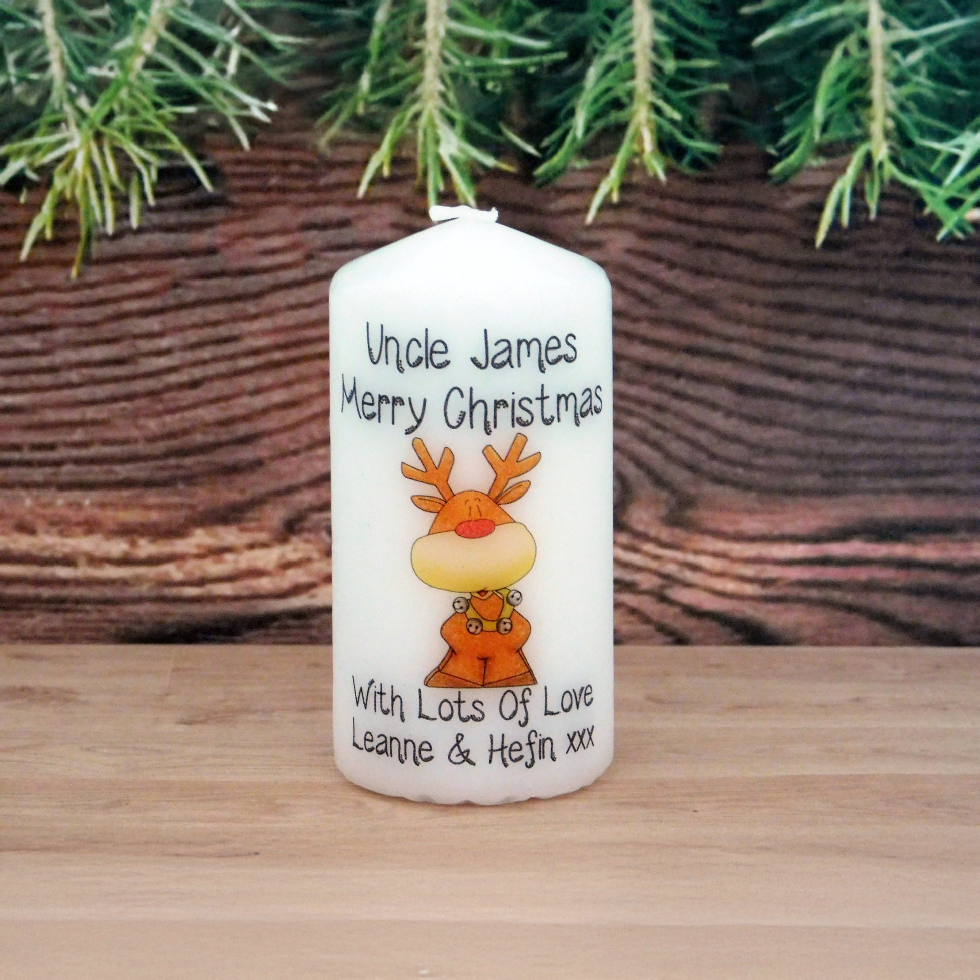 Personalized Christmas candle with a reindeer design and text, placed on a wooden surface with a wicker basket and greenery in the background.
