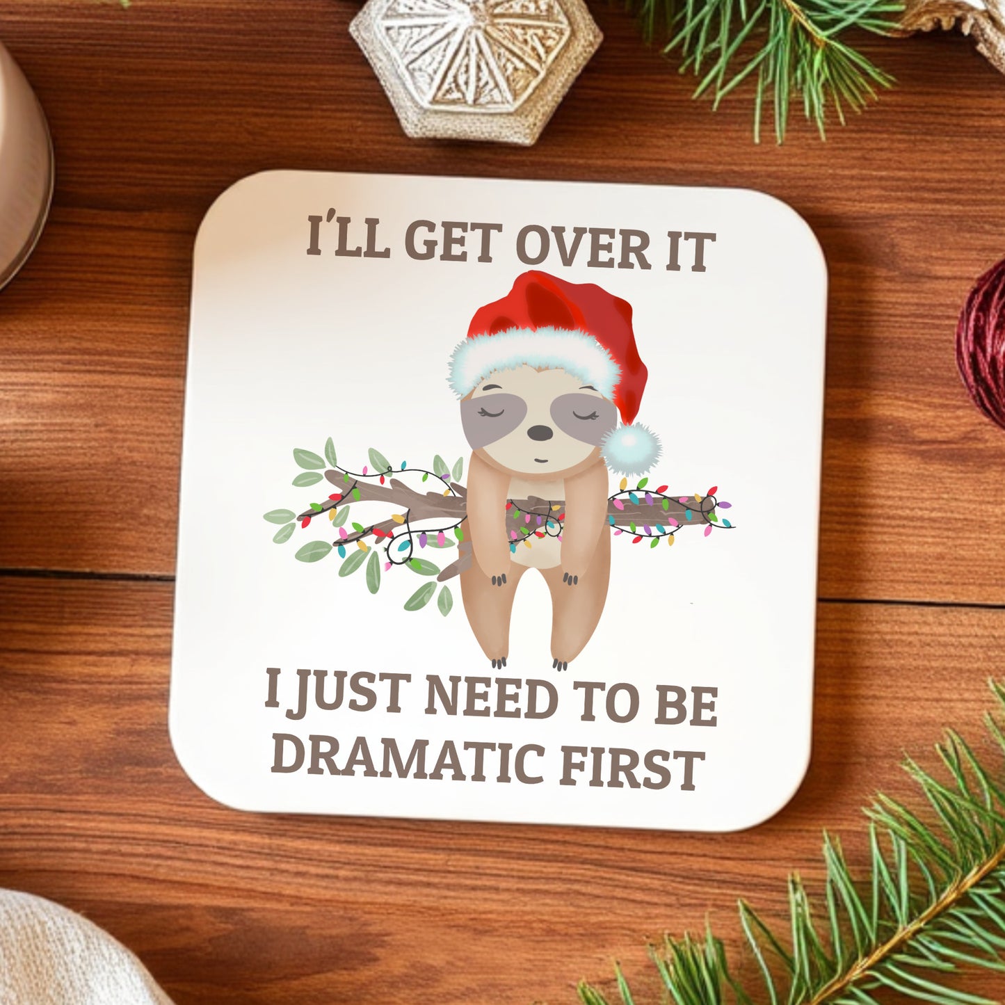 Coaster with sloth illustration and humorous text on a wooden surface with Christmas decorations.