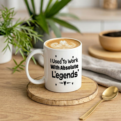 I Used To Work With Absolute Legends' white ceramic mug filled with latte art, displayed on a wooden board with a gold spoon and houseplants in the background