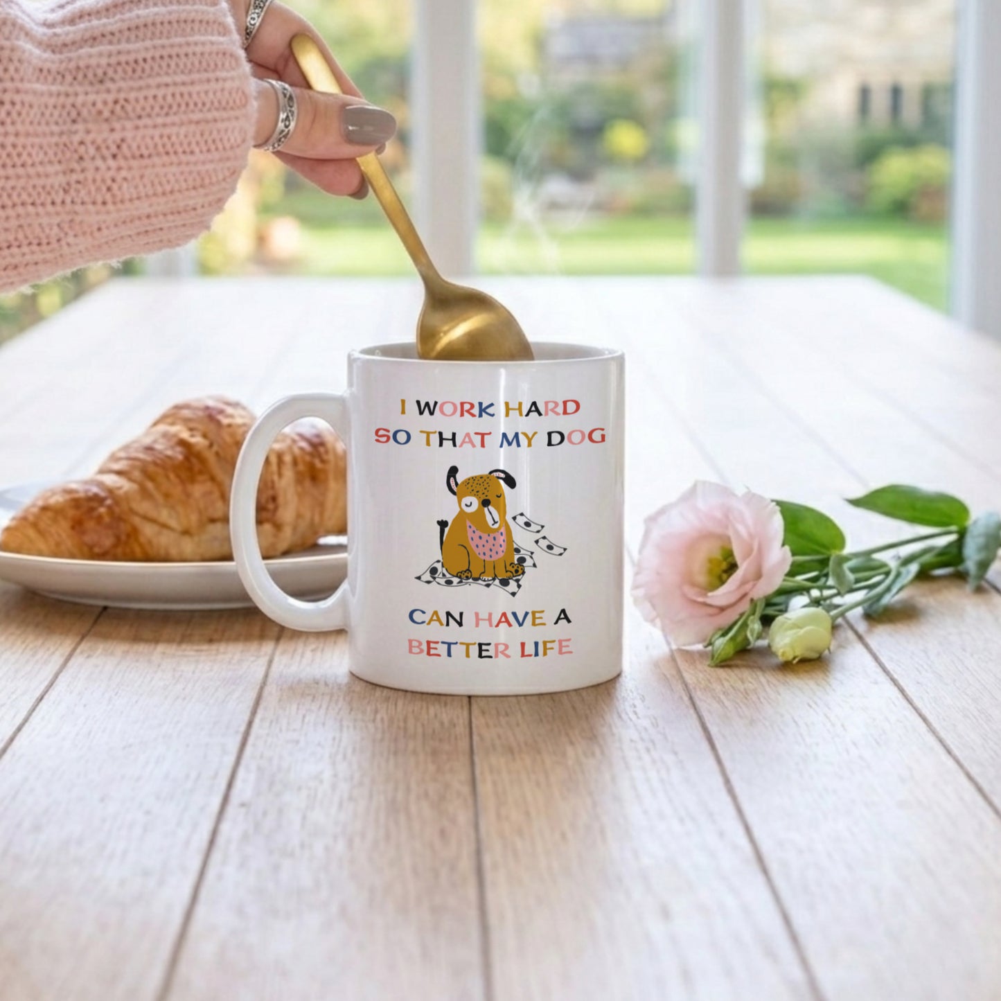 White ceramic mug with funny dog quote 'I Work Hard So That My Dog Can Have a Better Life' and cute illustrated dog with money, being stirred with a gold spoon on a wooden table with a croissant and pink rose
