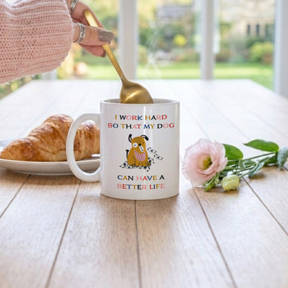 White ceramic mug with funny dog quote 'I Work Hard So That My Dog Can Have a Better Life' and cute illustrated dog with money, being stirred with a gold spoon on a wooden table with a croissant and pink rose