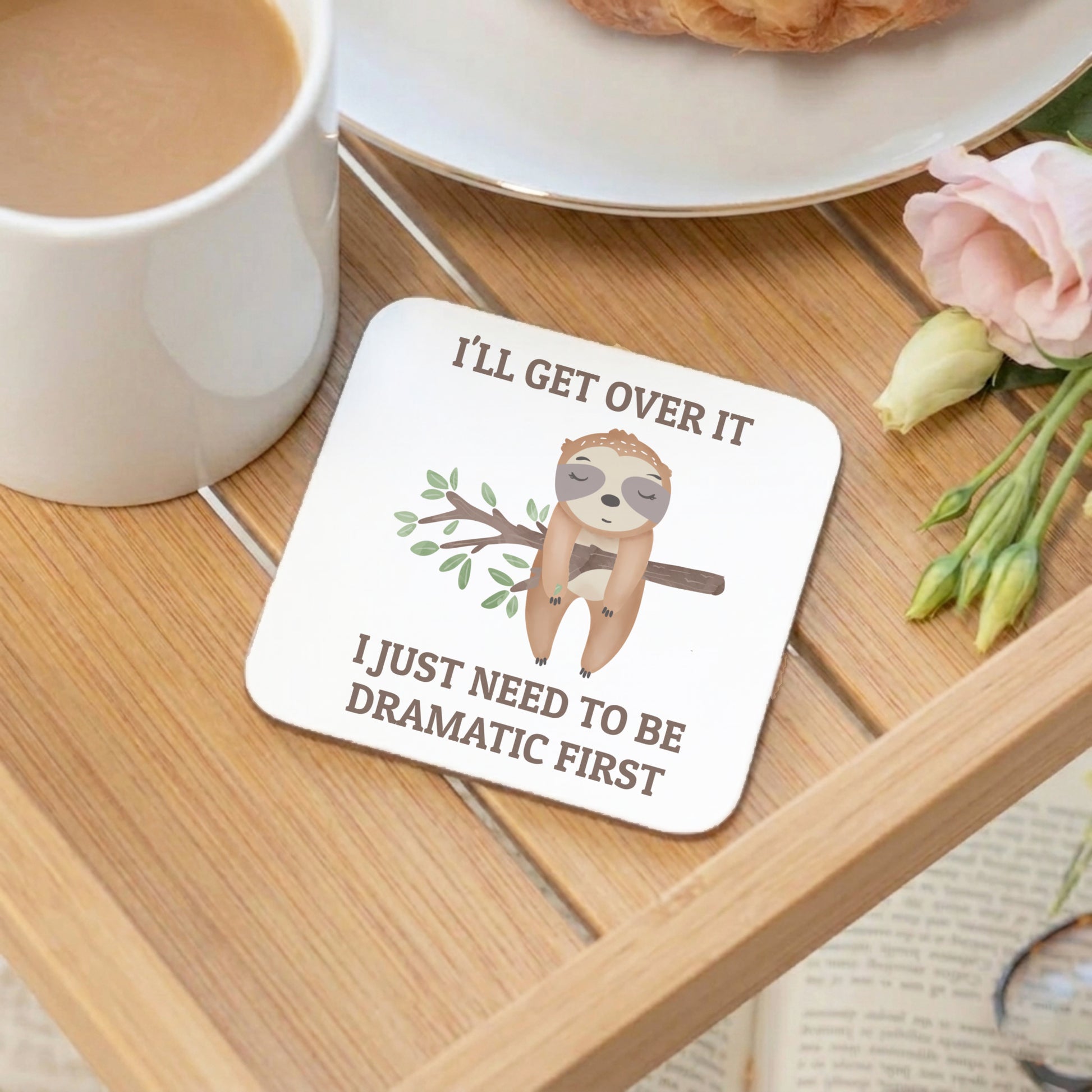 Coaster with sloth illustration and humorous text on a wooden table with coffee and flowers.