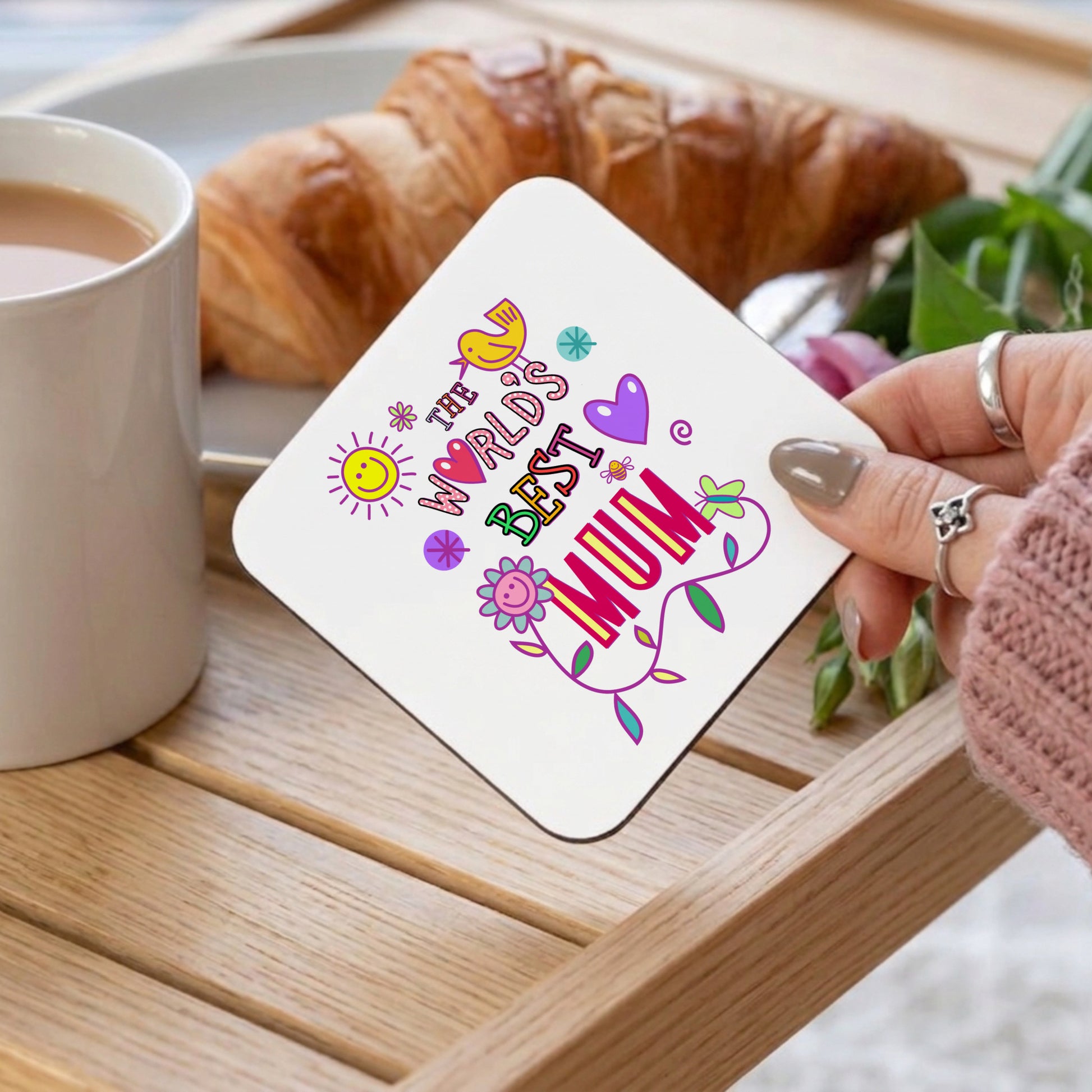Coaster with 'The World's Best Mum' text held by a hand over a cup of coffee and croissant on a wooden table.