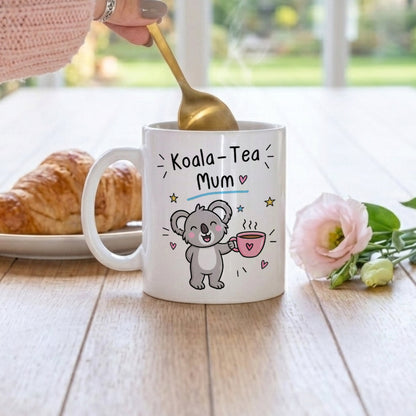 A white ceramic mug with a 'Koala-Tea Mum' cartoon graphic sits on a wooden table, with a hand holding a gold spoon above it. A croissant is on the left and a pink flower is on the right, set against a blurred outdoor background.