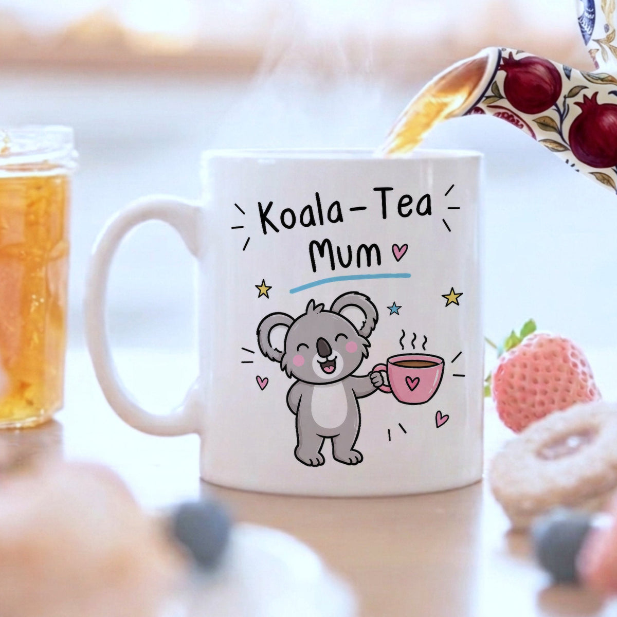 A white ceramic mug with a cartoon koala holding a pink teacup, printed with the text 'Koala-Tea Mum'. A patterned teapot is pouring tea into the mug, set on a table with a blurred breakfast scene including a strawberry, donuts, and a jar of iced tea.