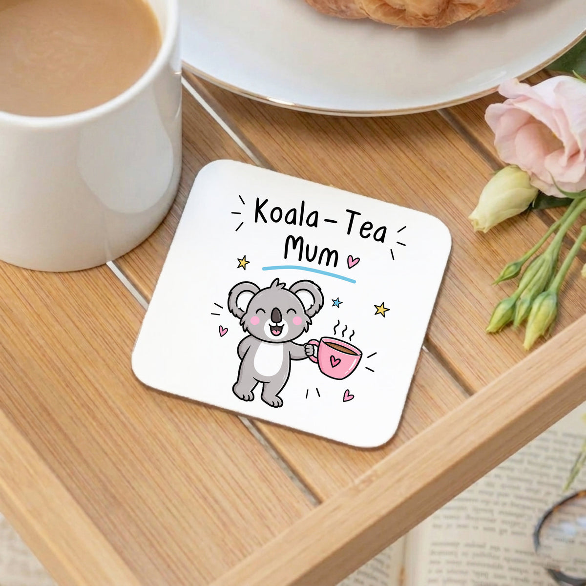 A square white coaster with rounded corners sits on a wooden table, printed with the text 'Koala-Tea Mum' above a cute cartoon koala holding a pink teacup. A white mug of coffee, a plate with a pastry, and pink flowers are arranged around it.