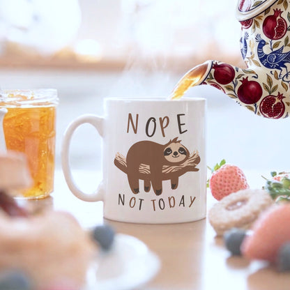 'Nope Not Today' sloth mug being filled from a floral teapot, displayed on a table with iced tea, strawberries, and blueberries