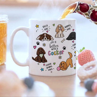 A white ceramic mug with a colourful cartoon dog print and the text 'Life is better with a Cocker' is shown on a tabletop with tea being poured into it, alongside a jar of honey, a strawberry, and blurred pastries in the background.
