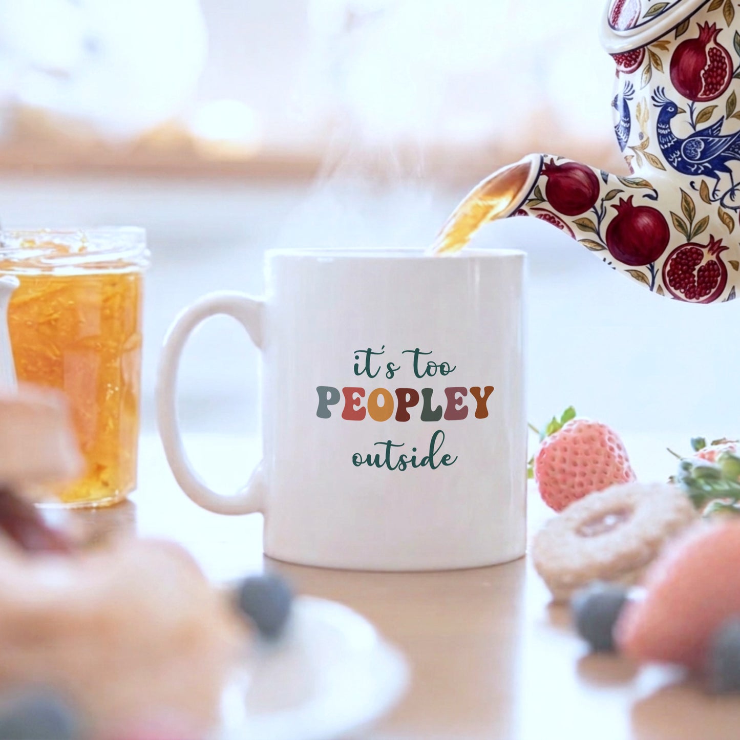 White mug with 'it's too peopley outside' text, being filled with tea, on a blurred background of food and drinks.