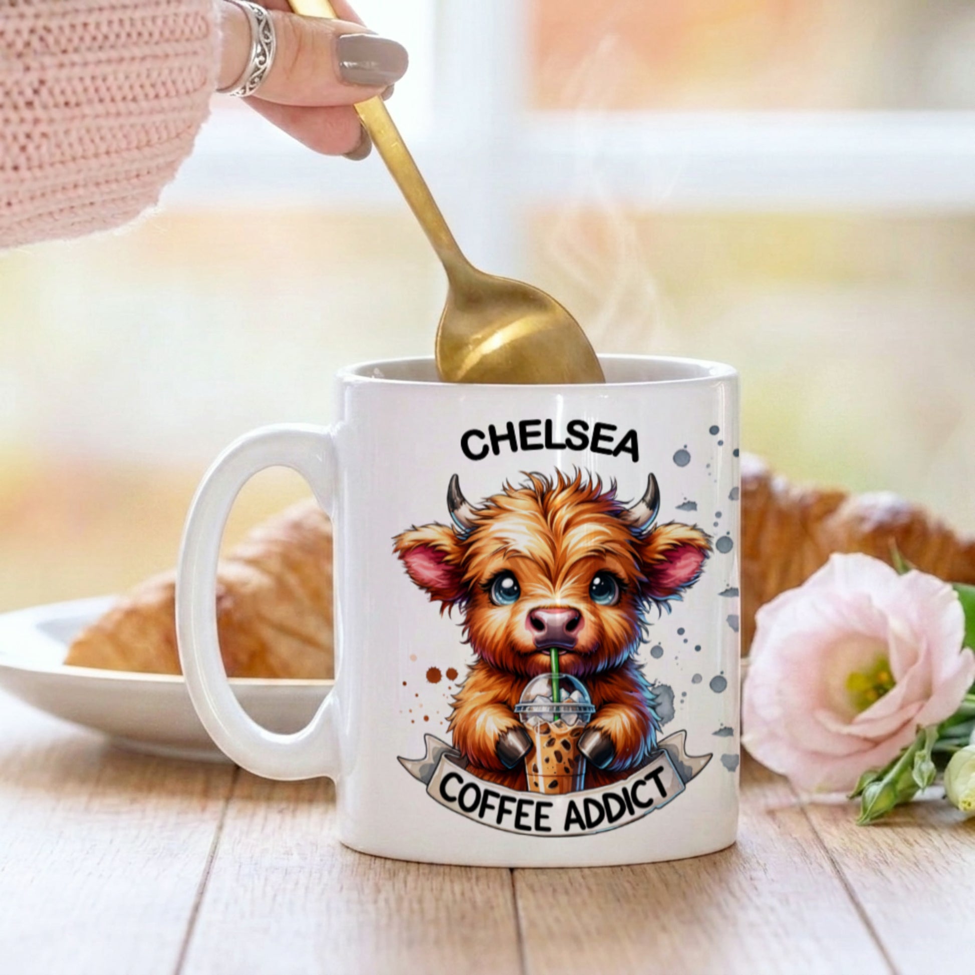 Mug with a cartoon cow design and 'Chelsea Coffee Addict' text, held by a hand with a spoon.