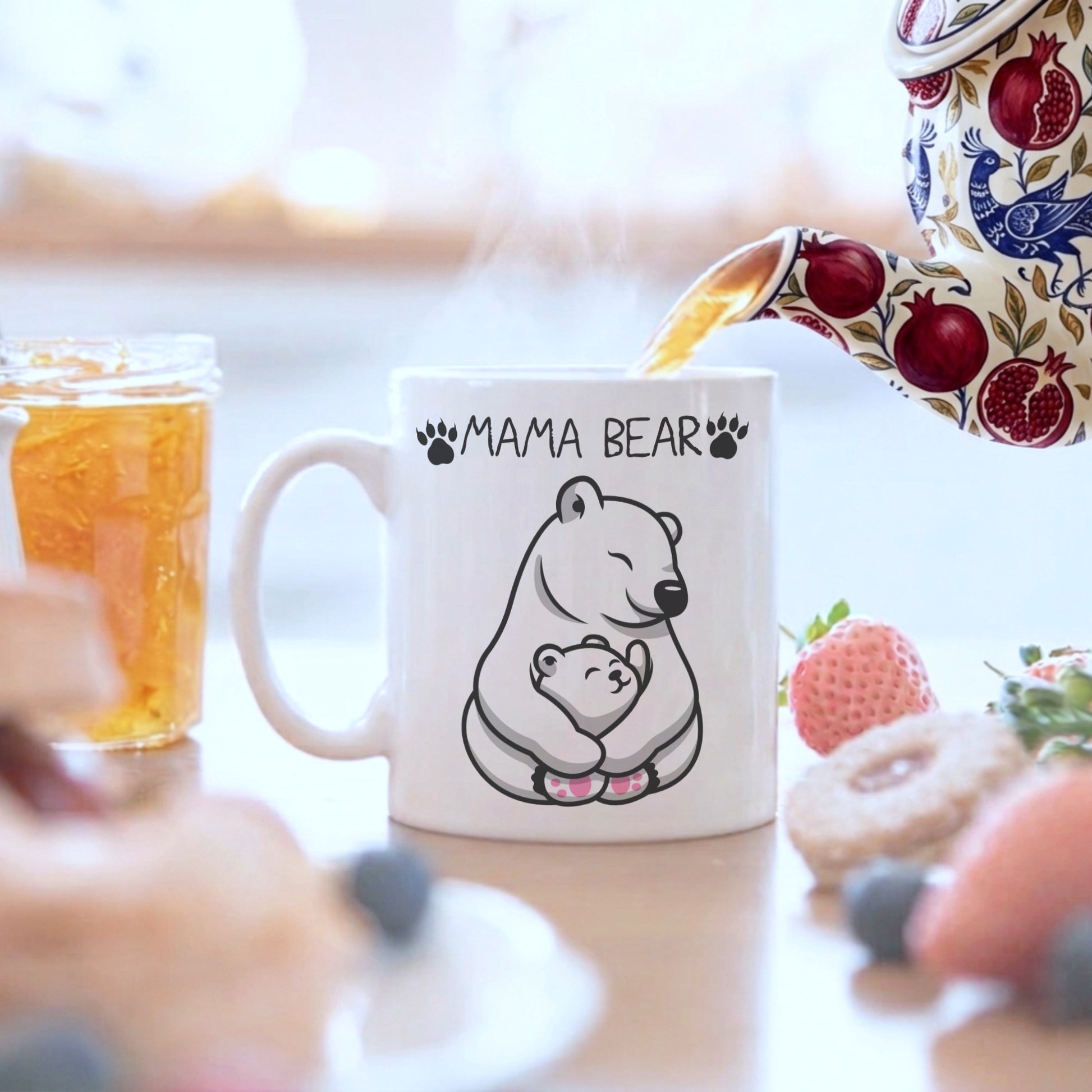 A white ceramic mug with a black line art illustration of a mother bear and cub, with the text 'MAMA BEAR' above. A floral teapot is pouring tea into the mug, which is set on a table with breakfast items like a jar of honey, a strawberry, and donuts in a blurred kitchen setting.