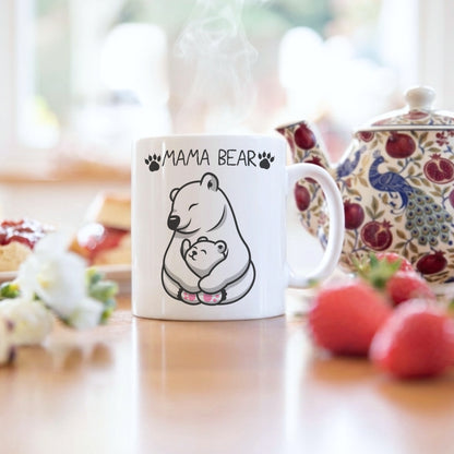 A white ceramic mug with a black line art illustration of a mama bear holding her cub, with the text 'MAMA BEAR' above. The mug is shown on a wooden table with a teapot and strawberries in a warm, blurred kitchen setting.