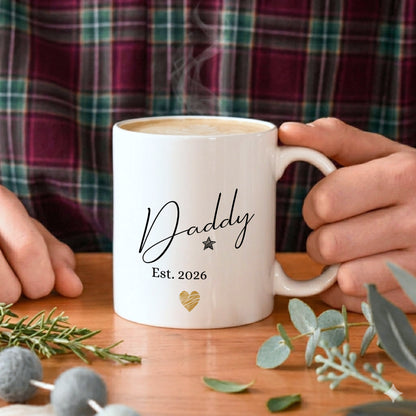 White mug with 'Daddy Est. 2026' text held by a person, with a plaid shirt in the background.