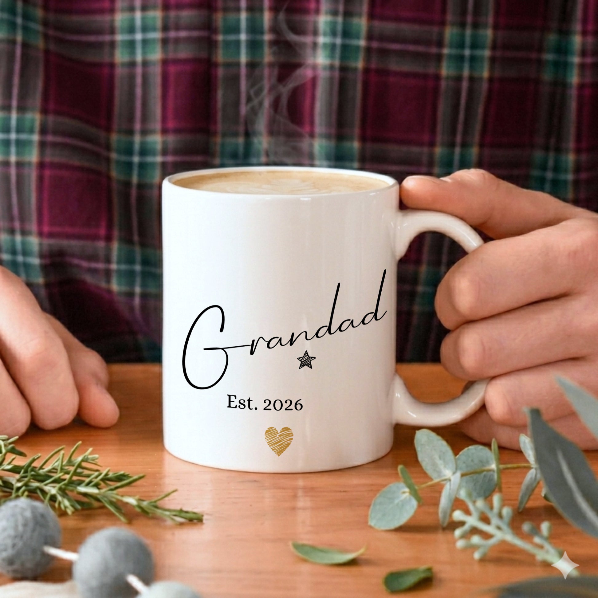 White mug with 'Grandad Est. 2026' text held by a person, with a plaid shirt in the background.