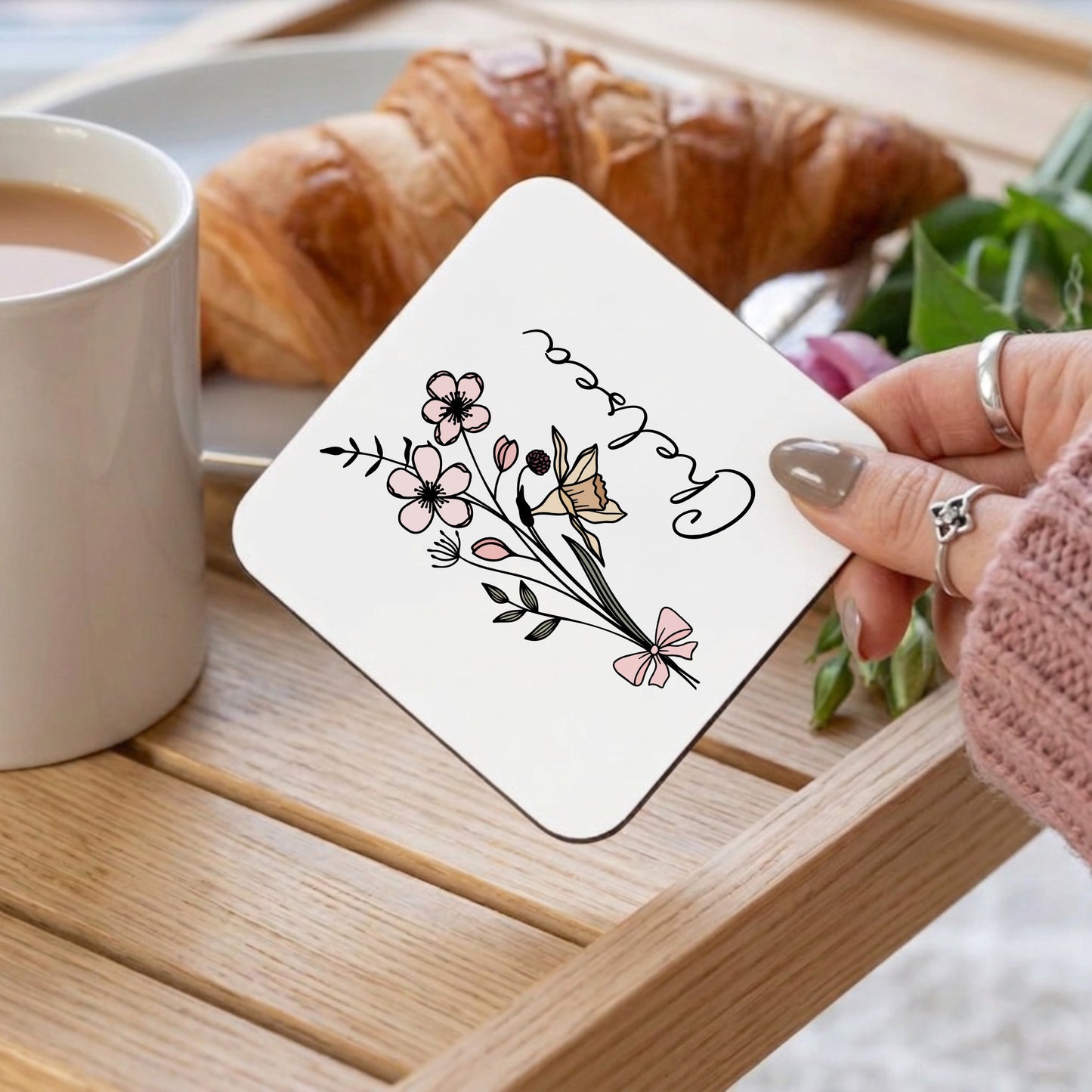Coaster with floral design held by a hand over a cup of coffee and croissant on a wooden table.