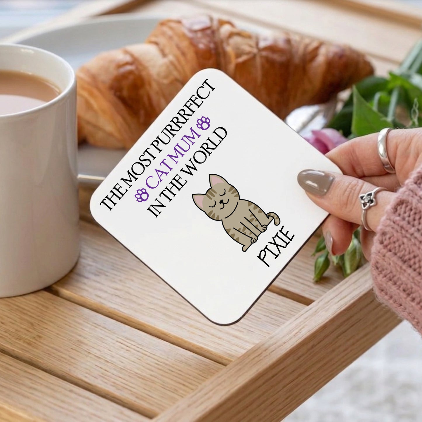 Woman holding a coaster with cat illustrations and above a tray with food and tea.