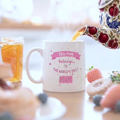 A white mug which says this mug belongs to the world's best mum on a breakfast table with a teapot pouring tea