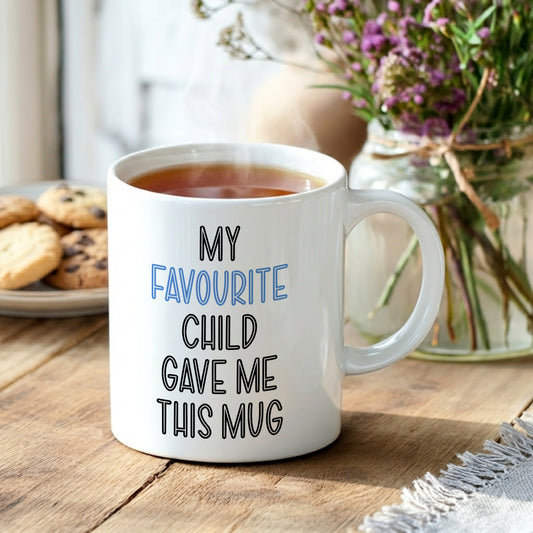 White mug with text 'My favourite child gave me this mug' on a wooden table with tea and cookies.