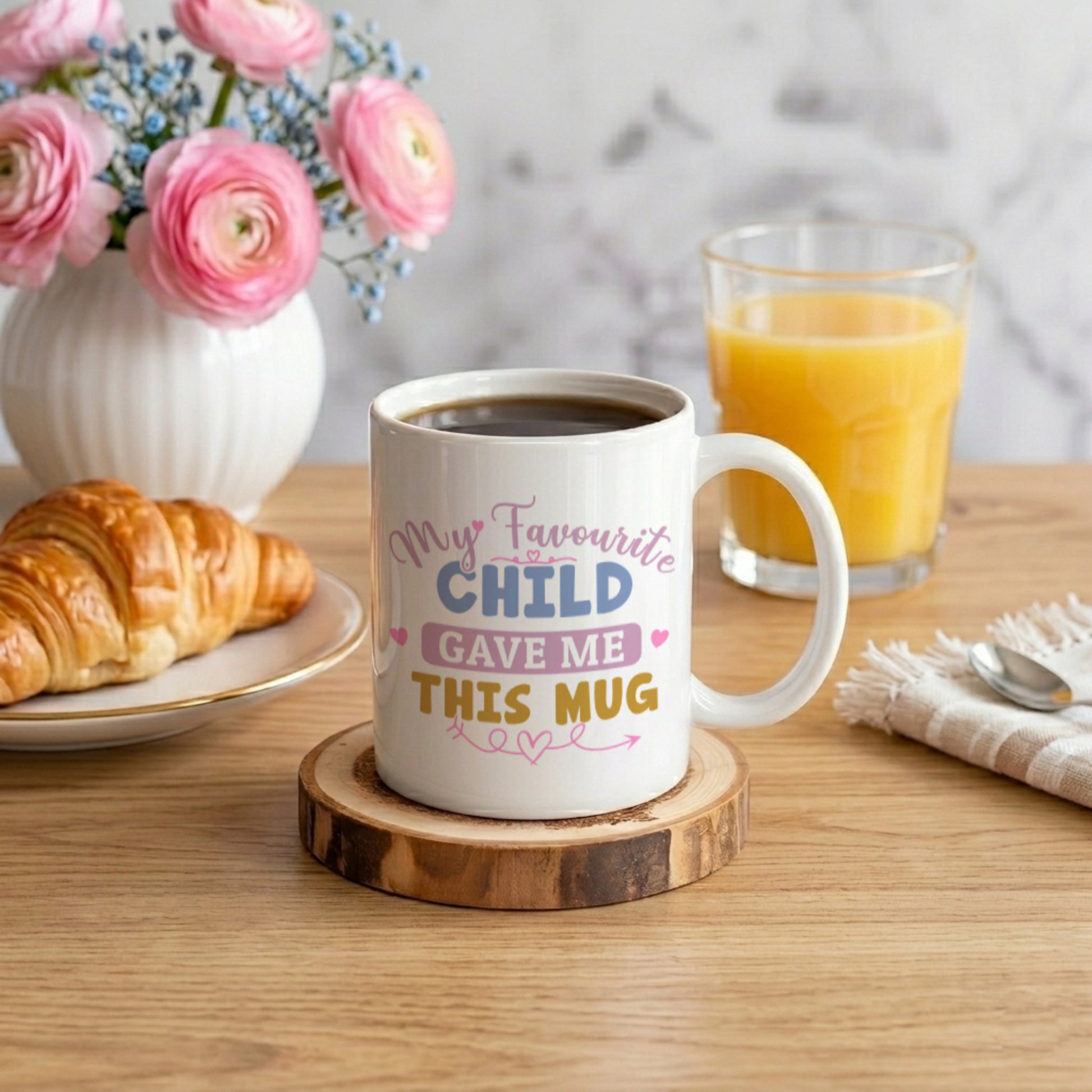 Mug with 'My favourite child gave me this mug' text on a wooden coaster with coffee, croissant, and juice.