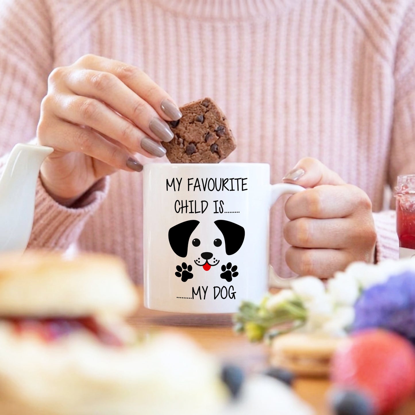 Person holding a mug with a dog illustration and text, surrounded by food and a blurred background
