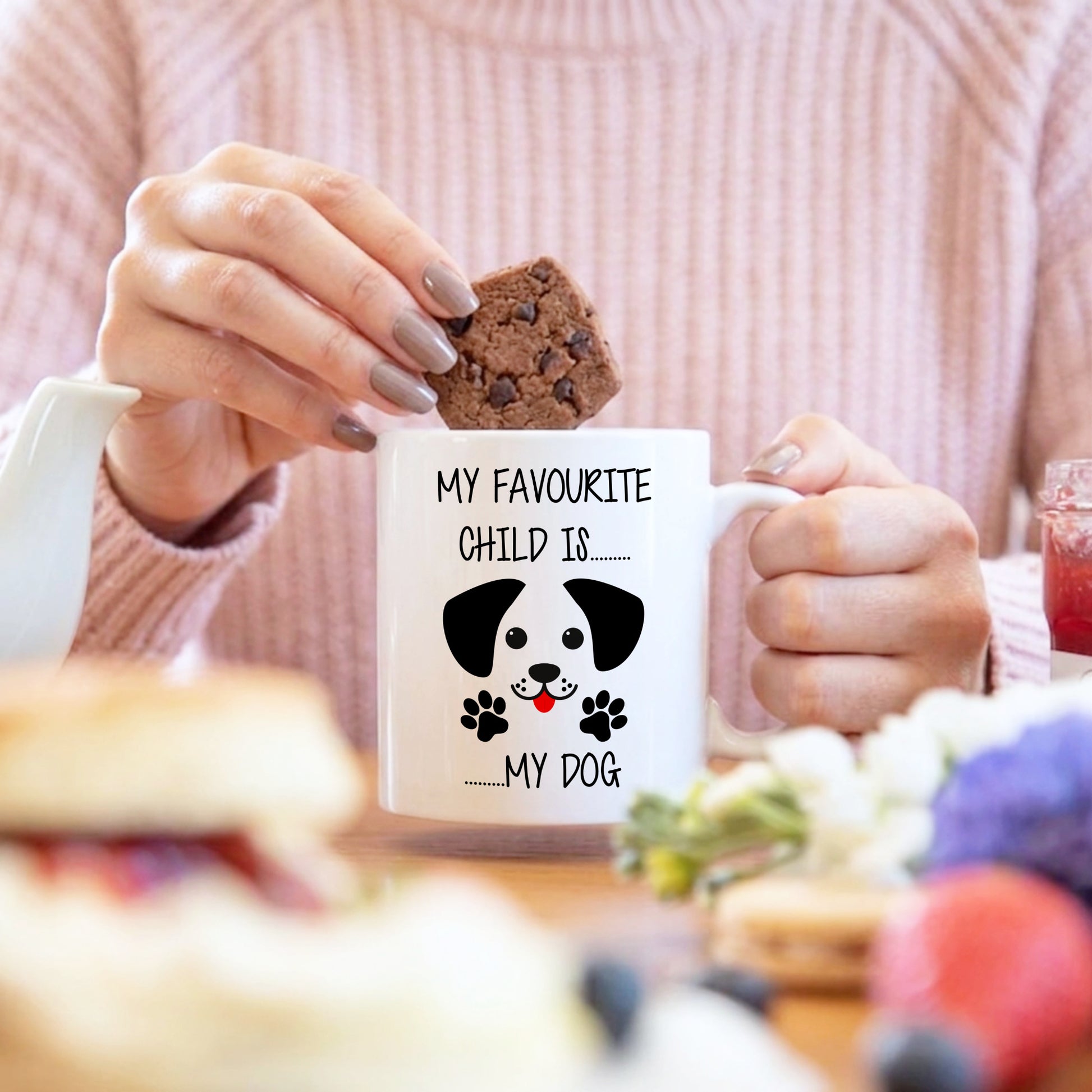 Person holding a mug with a dog illustration and text, surrounded by food and a blurred background