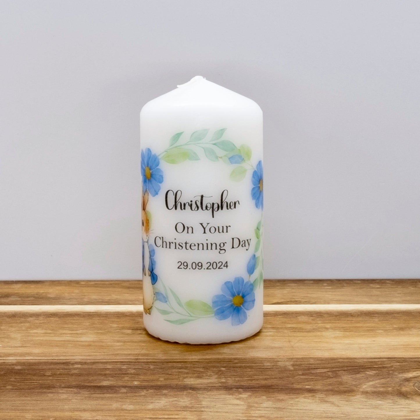 A personalised white pillar candle with blue flowers and green leaves, printed with the name 'Christopher', 'On Your Christening Day', and the date '29.09.