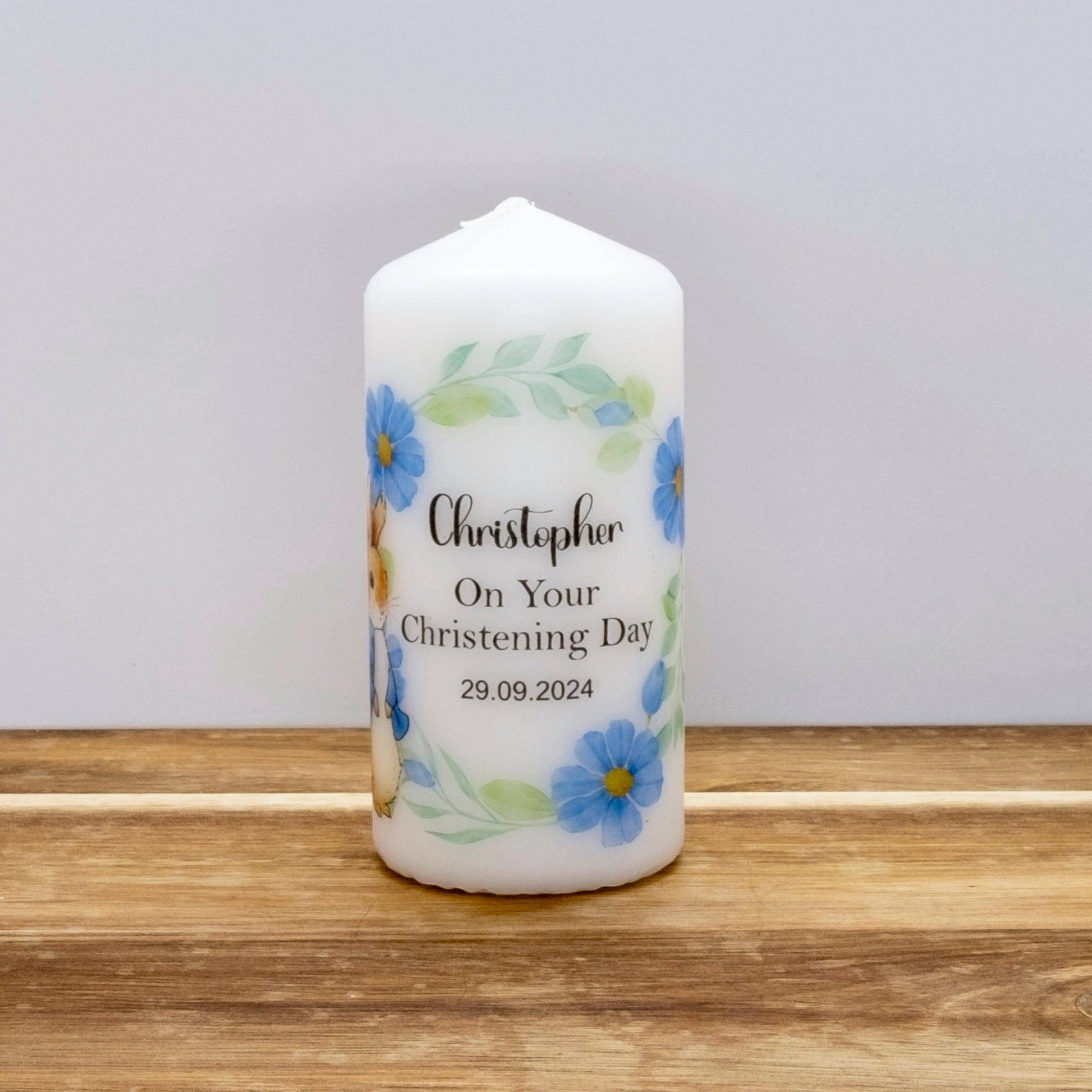 A personalised white pillar candle with blue flowers and green leaves, printed with the name 'Christopher', 'On Your Christening Day', and the date '29.09.