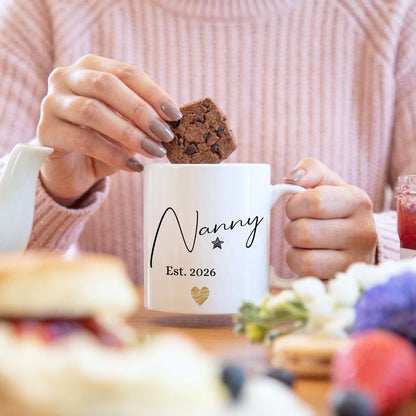 Woman dunking a chocolate brownie into a personalised 'Nanny Est. 2026' mug with a gold heart design, held with both hands