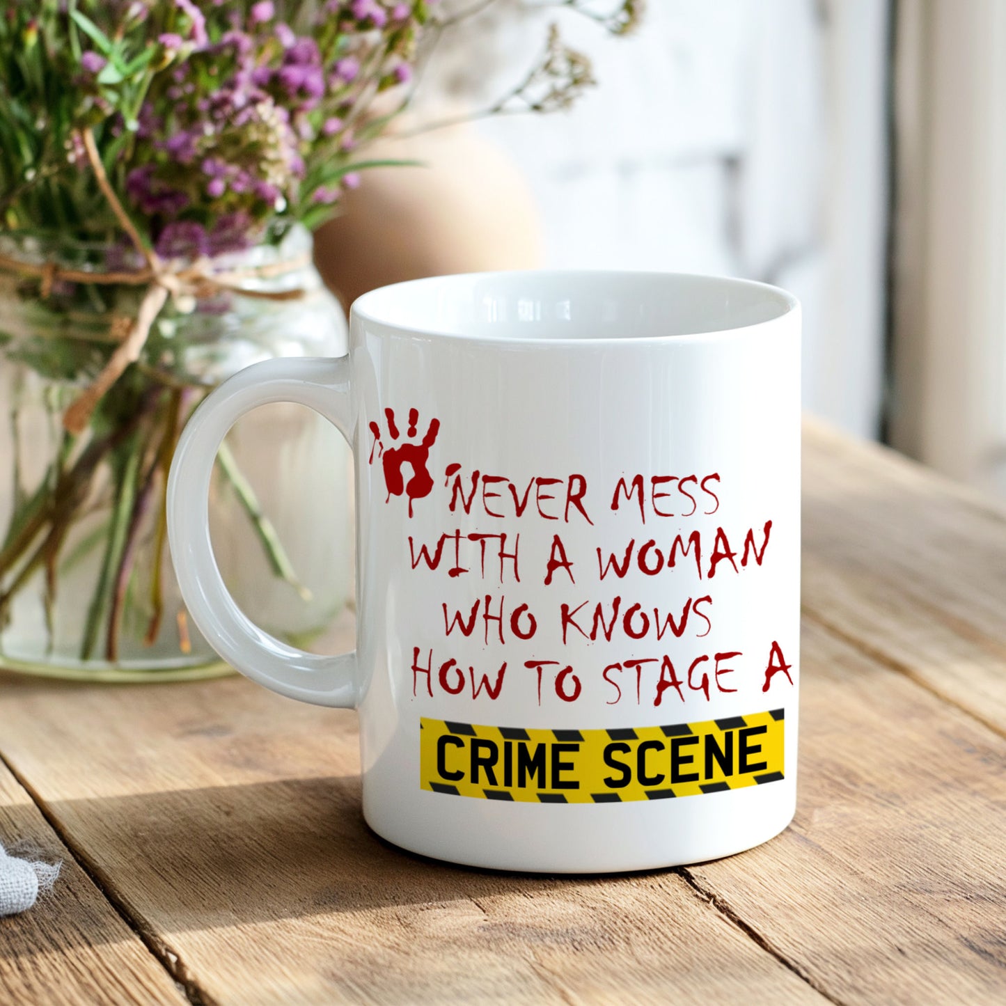 A white ceramic mug with a humorous crime scene quote printed in red and yellow, placed on a rustic wooden table with a blurred vase of purple flowers in the background.