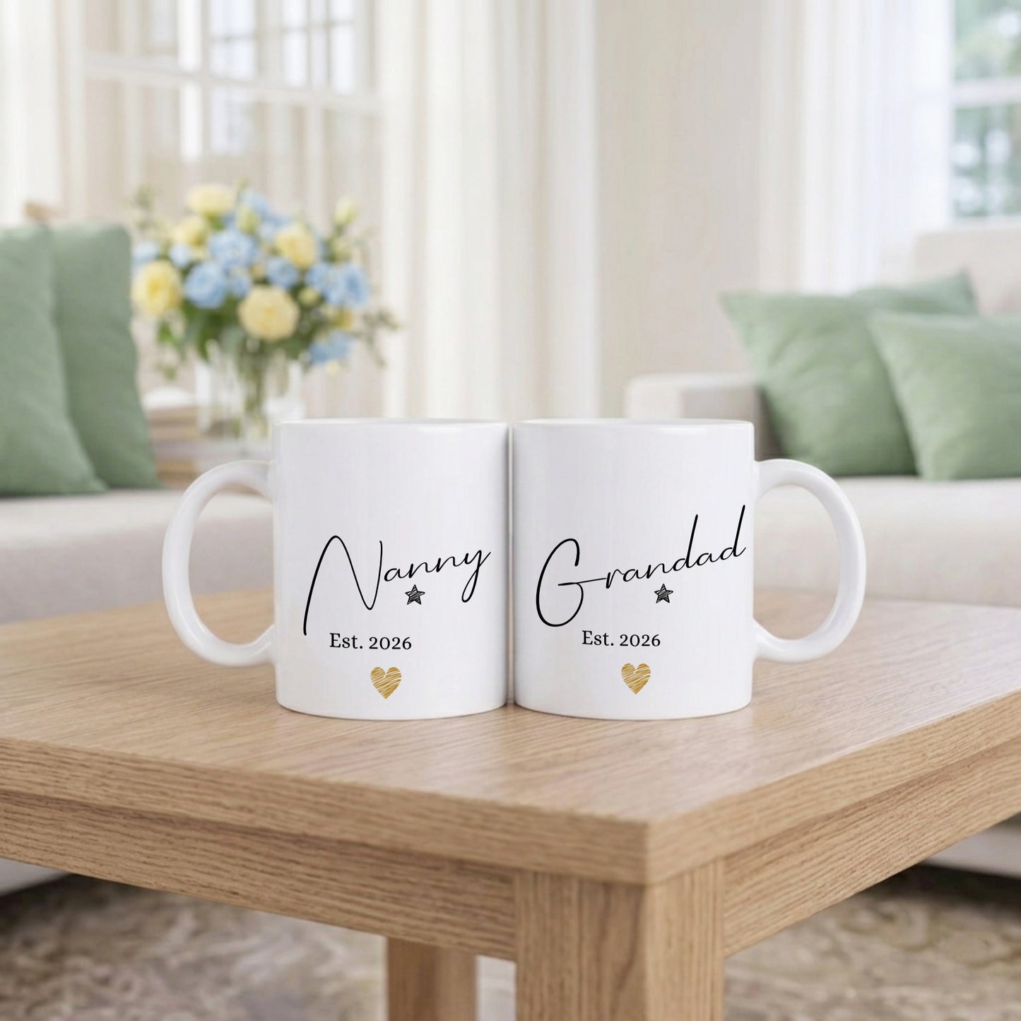 Two white mugs with 'Nanny Est. 2026' and 'Grandad Est. 2026' text on a wooden table in a living room setting.