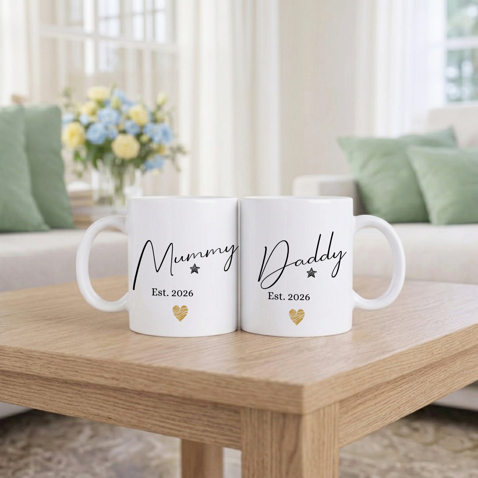 Two white mugs with 'Mummy Est. 2026' and 'Daddy Est. 2026' text on a wooden table in a living room setting.