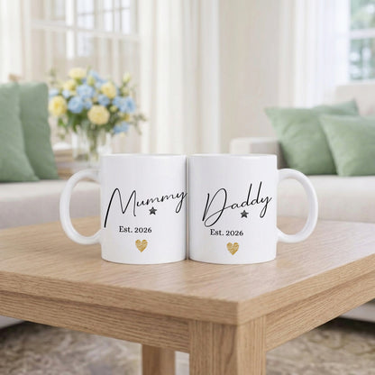Two white mugs with 'Mummy Est. 2026' and 'Daddy Est. 2026' text on a wooden table in a living room setting.