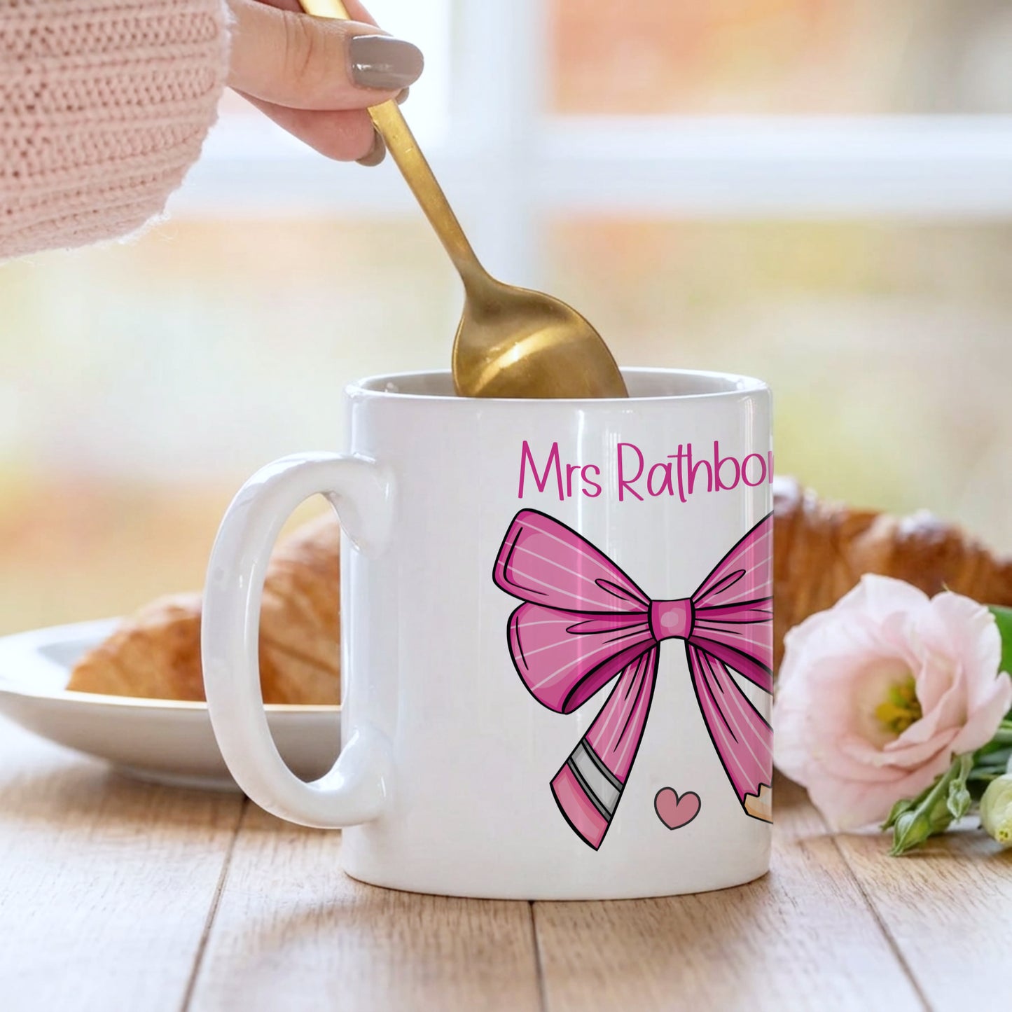 Personalised teacher mug with a pink bow and pencil design customised with 'Mrs Rathbone', being stirred with a gold spoon alongside a croissant and pink flower