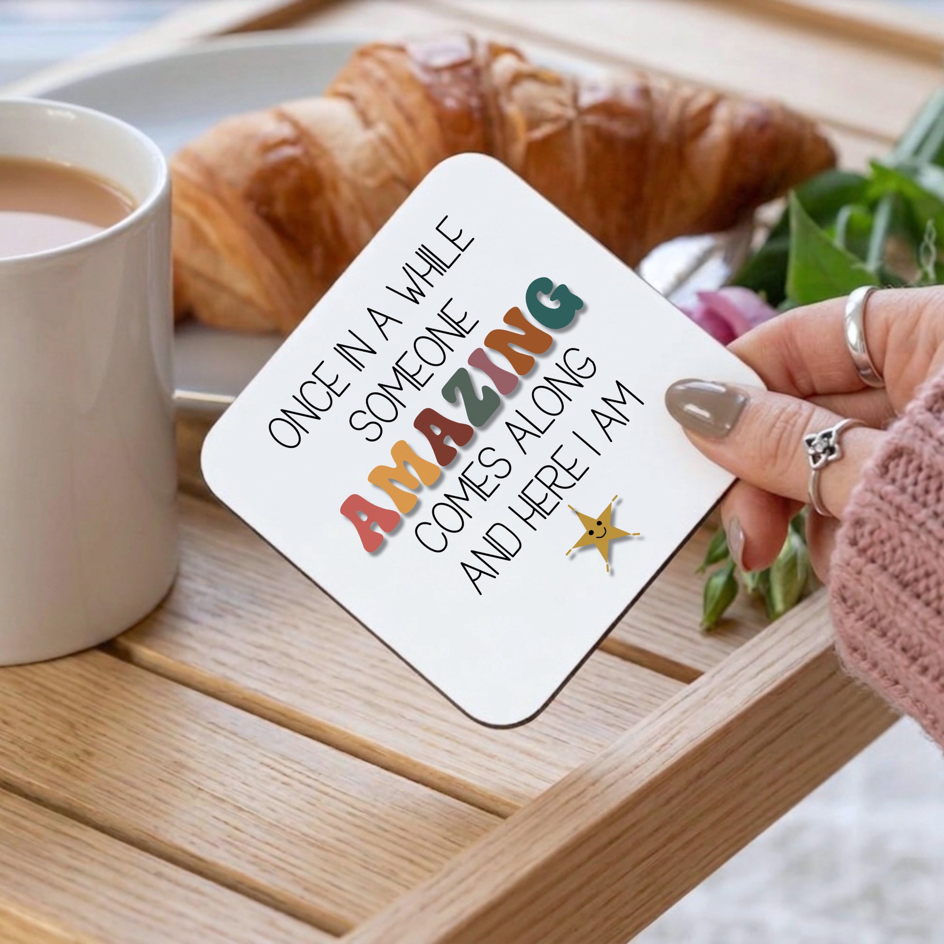 Coaster with motivational quote held by a hand on a table with coffee and croissant