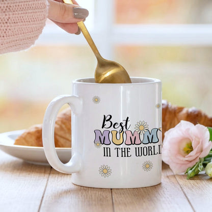 White mug with 'Best Mummy in the World' text, held by a hand with a gold spoon, on a wooden table with a blurred background.
