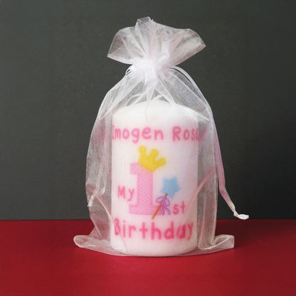 A white candle wrapped in a sheer white organza bag, printed with pink text reading 'Imogen Rose My 1st Birthday' and decorated with a pink number one, yellow crown, and blue star wand, set against a dark grey background and red surface.