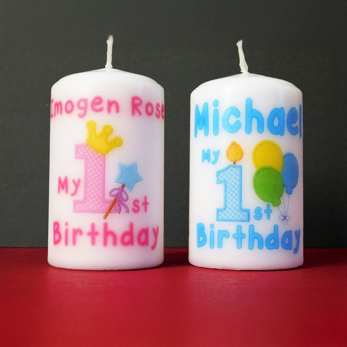 Two personalised white birthday candles standing upright on a red surface against a dark grey background. The left candle reads 'Imogen Rose My 1st Birthday' with pink text, a pink number 1, a yellow crown, and a blue star.