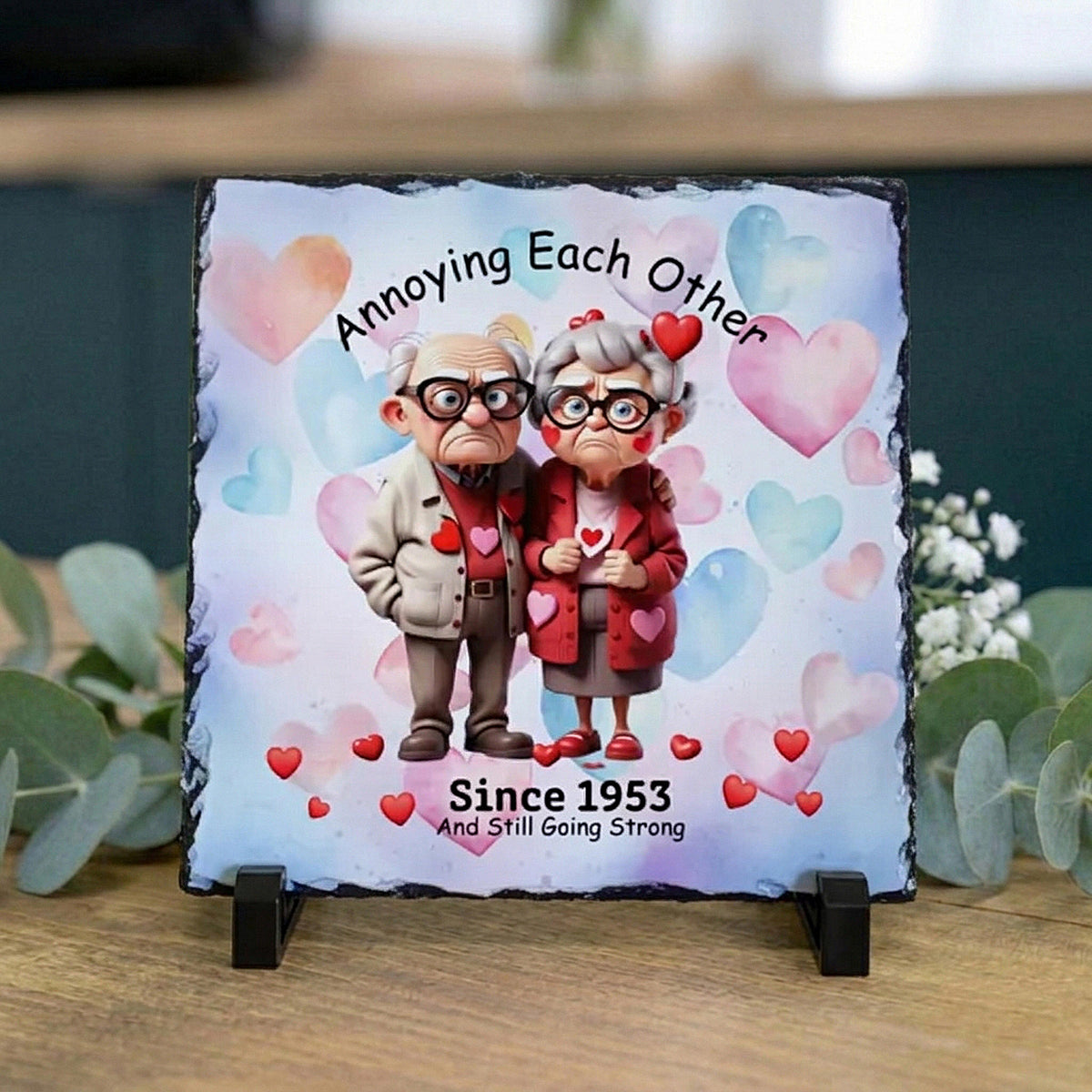 A square slate sign on a small black stand, featuring a custom cartoon illustration of an elderly couple with hearts and the text 'Annoying Each Other Since 1953 And Still Going Strong.' The sign is displayed on a wooden surface with eucalyptus leaves in the background.
