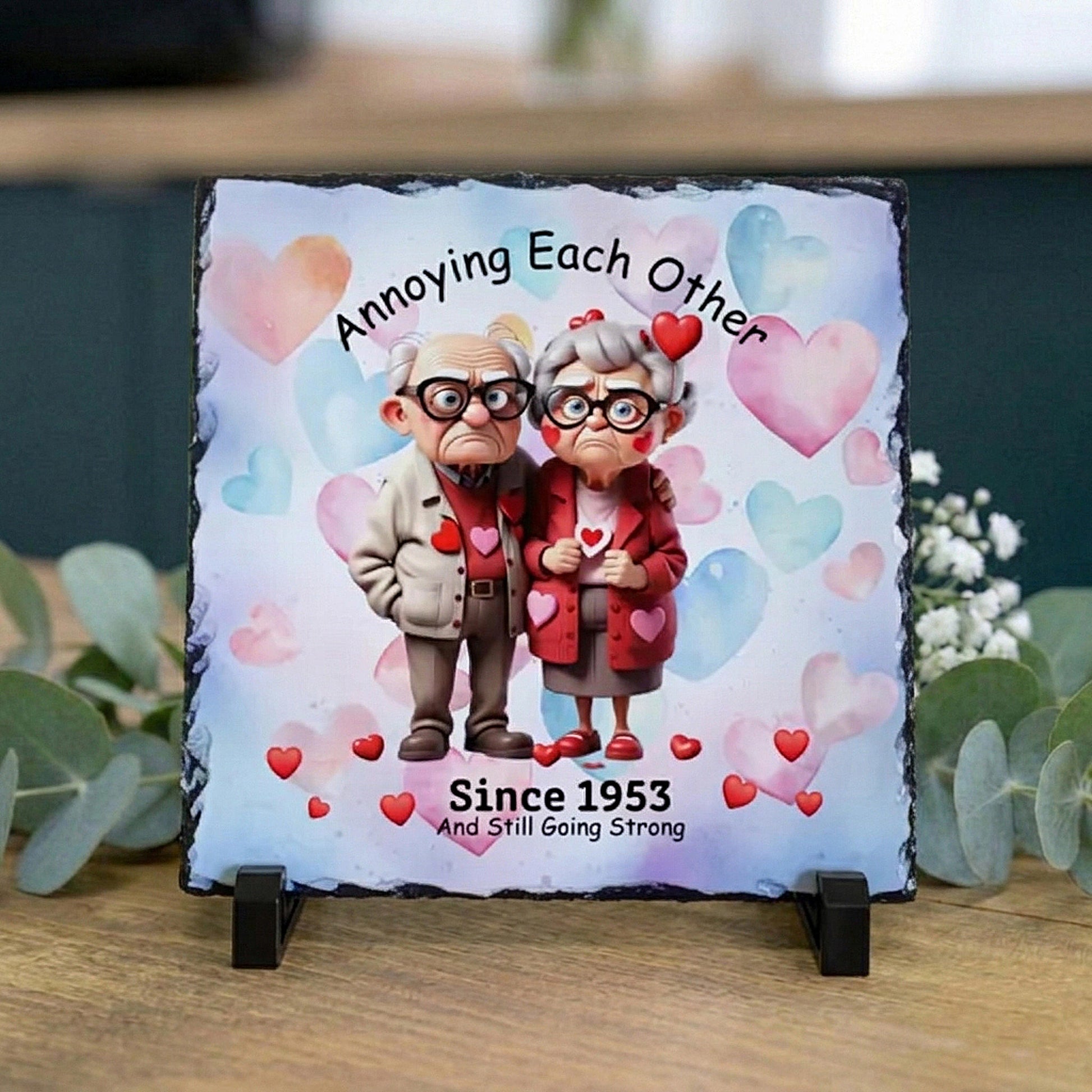A square slate sign on a small black stand, featuring a custom cartoon illustration of an elderly couple with hearts and the text 'Annoying Each Other Since 1953 And Still Going Strong.' The sign is displayed on a wooden surface with eucalyptus leaves in the background.