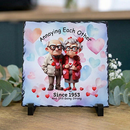 A square slate sign on a small black stand, featuring a custom cartoon illustration of an elderly couple with hearts and the text 'Annoying Each Other Since 1953 And Still Going Strong.' The sign is displayed on a wooden surface with eucalyptus leaves in the background.