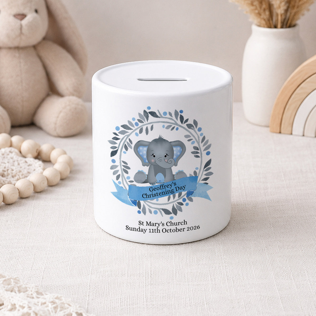 Two white ceramic baby savings jars with slot tops, decorated with cute gray elephant illustrations and floral wreaths. One jar has a blue ribbon banner for 'Bethany's First Communion' and the other has a pink ribbon banner for 'Ella's First Holy Communion'.