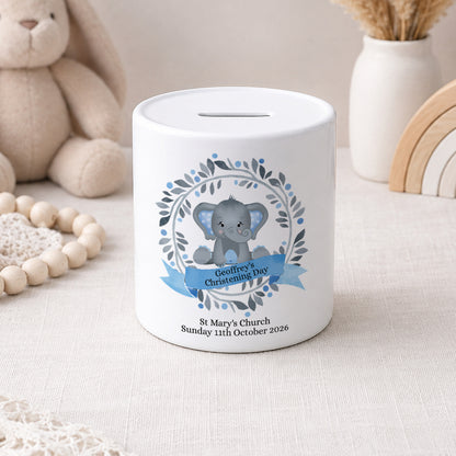 Two white ceramic baby savings jars with slot tops, decorated with cute gray elephant illustrations and floral wreaths. One jar has a blue ribbon banner for 'Bethany's First Communion' and the other has a pink ribbon banner for 'Ella's First Holy Communion'.