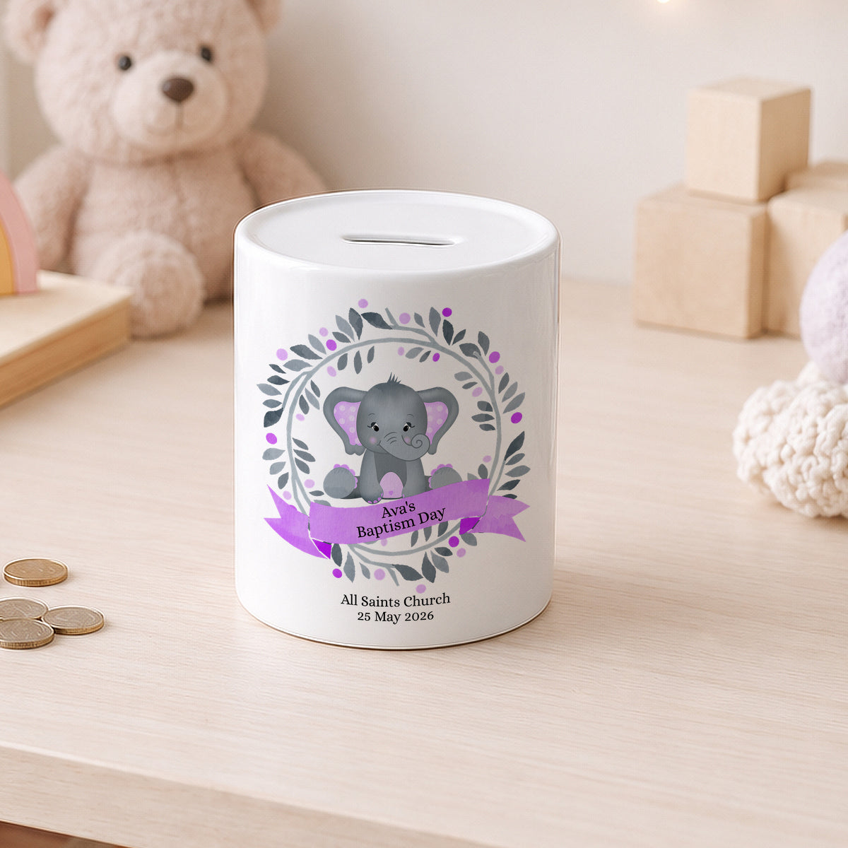 A white ceramic baby savings bank with a grey elephant illustration and purple ribbon, set on a light wooden table with a teddy bear in the background.