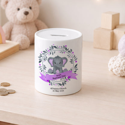 A white ceramic baby savings bank with a grey elephant illustration and purple ribbon, set on a light wooden table with a teddy bear in the background.