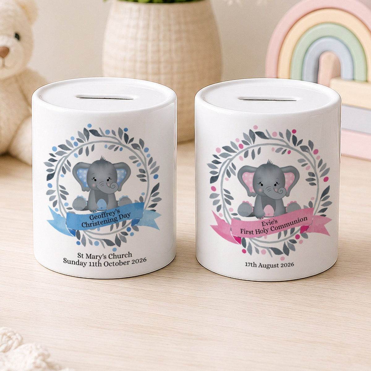 Two white ceramic baby savings jars with slot tops, decorated with cute gray elephant illustrations and floral wreaths. One jar has a blue ribbon banner for 'Bethany's First Communion' and the other has a pink ribbon banner for 'Ella's First Holy Communion'.