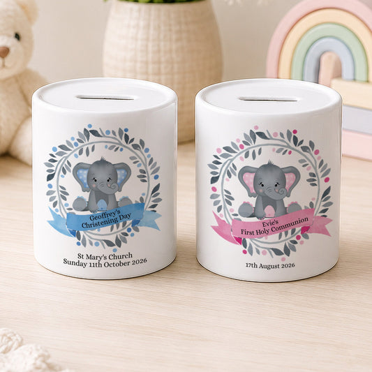Two white ceramic baby savings jars with slot tops, decorated with cute gray elephant illustrations and floral wreaths. One jar has a blue ribbon banner for 'Bethany's First Communion' and the other has a pink ribbon banner for 'Ella's First Holy Communion'.