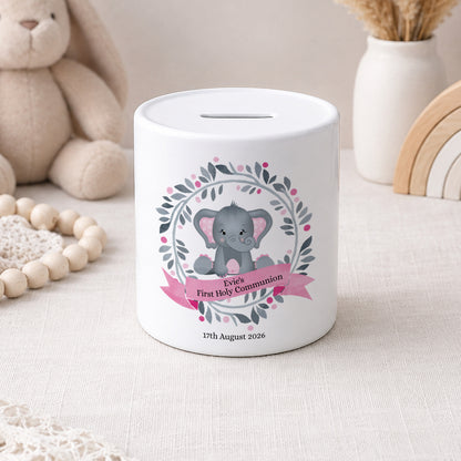 Two white ceramic baby savings jars with slot tops, decorated with cute gray elephant illustrations and floral wreaths. One jar has a blue ribbon banner for 'Bethany's First Communion' and the other has a pink ribbon banner for 'Ella's First Holy Communion'.