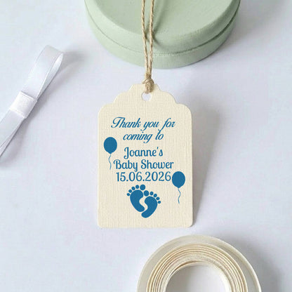A personalised baby shower gift tag with a blue printed design and text, hanging from a twine string. The tag is cream-coloured with blue balloon and baby footprint graphics.