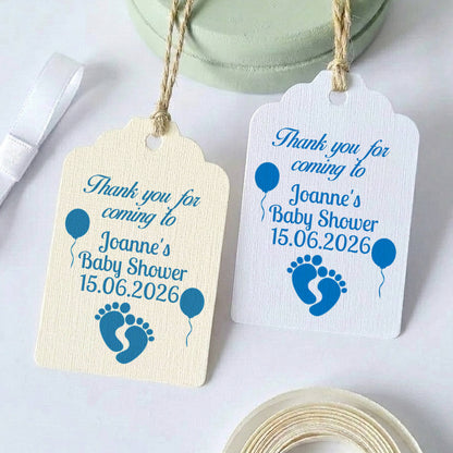 Two personalised baby shower gift tags with blue text and baby footprint/balloon graphics, shown on a white background with ribbon and tape props.
